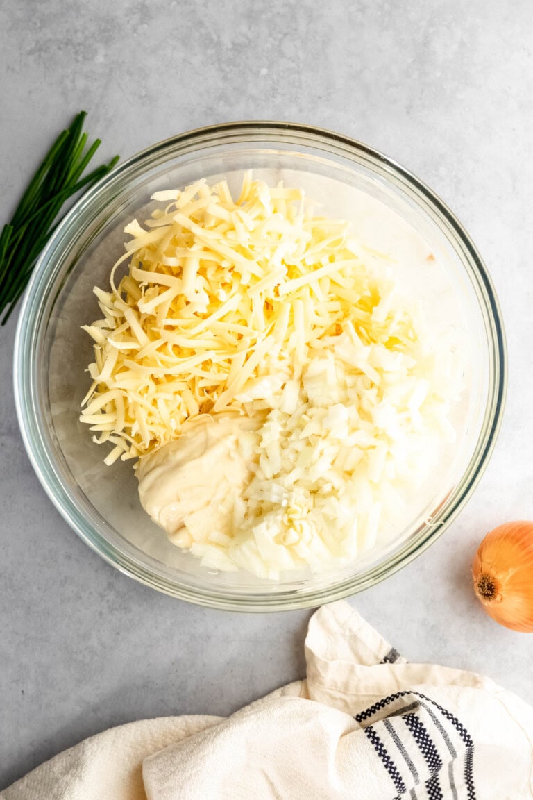 A glass bowl containing shredded cheese, chopped onions, and a dollop of mayonnaise on a light grey surface with chives, an onion, and a striped cloth nearby.