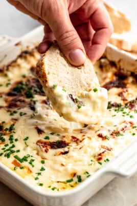 A hand dips a slice of bread into a creamy, baked cheese dip garnished with chopped chives in a white baking dish.