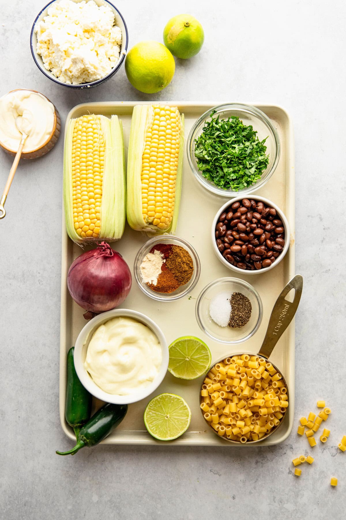 A tray holding ingredients including two ears of corn, pasta, black beans, chopped herbs, lime, red onion, jalapeños, spices, mayonnaise, and crumbled cheese.