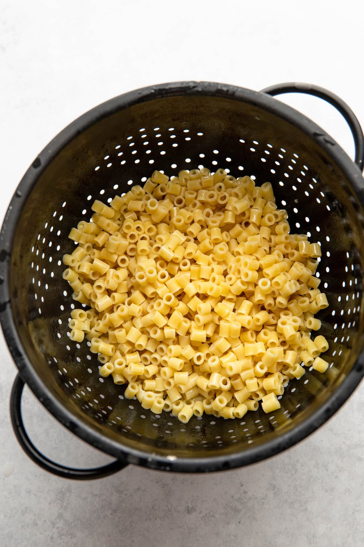 A black colander holds freshly cooked Ditalini pasta against a white background.