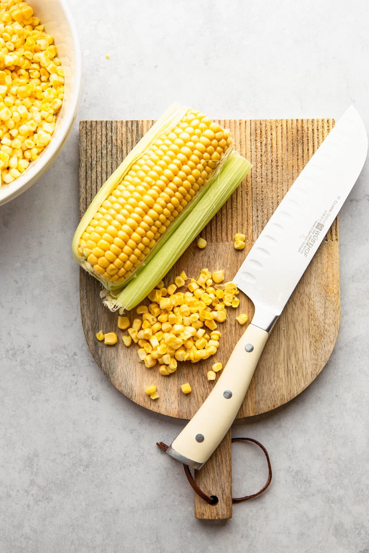 A cutting board with corn kernels, a partly husked corn cob, and a knife on it. A bowl with more corn kernels is in the background.