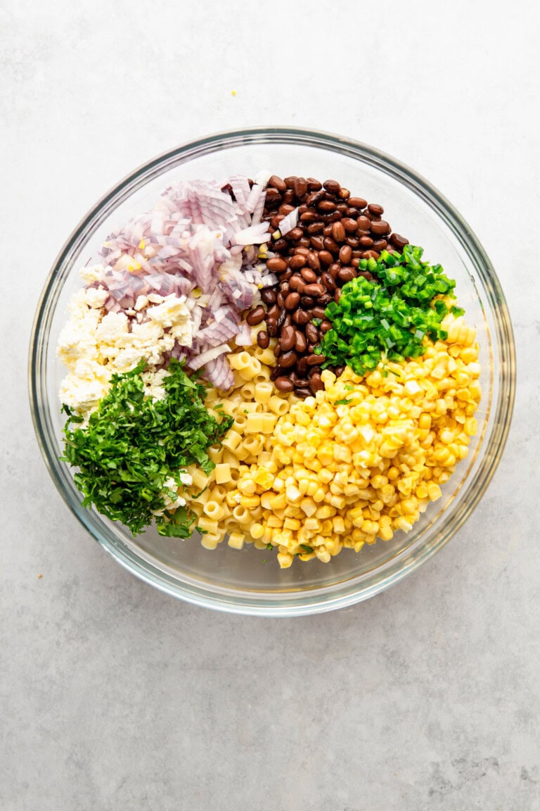 A glass bowl containing diced red onions, black beans, minced garlic, chopped green herbs, corn kernels, and cooked pasta on a gray surface.