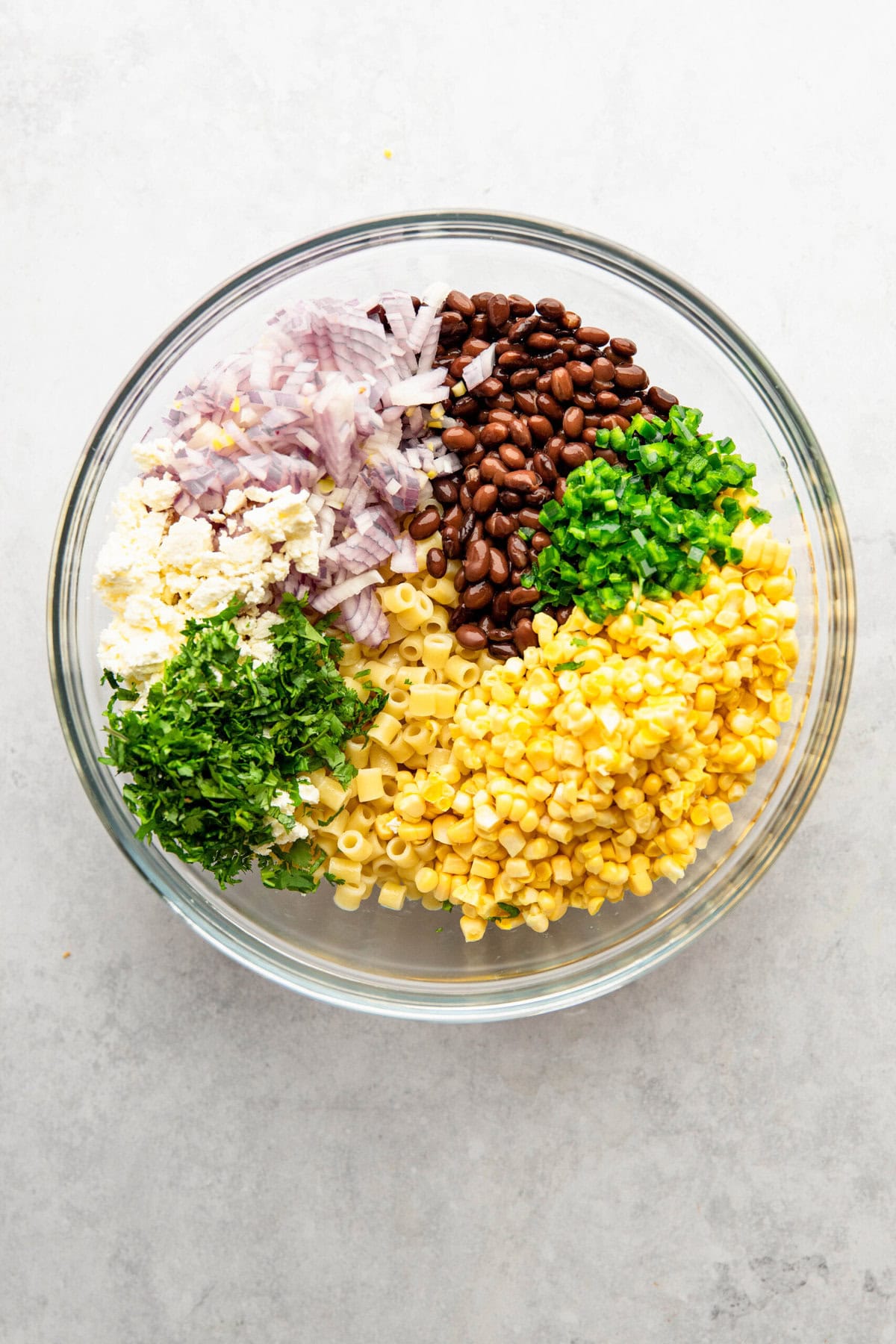 A glass bowl containing diced red onions, black beans, minced garlic, chopped green herbs, corn kernels, and cooked pasta on a gray surface.