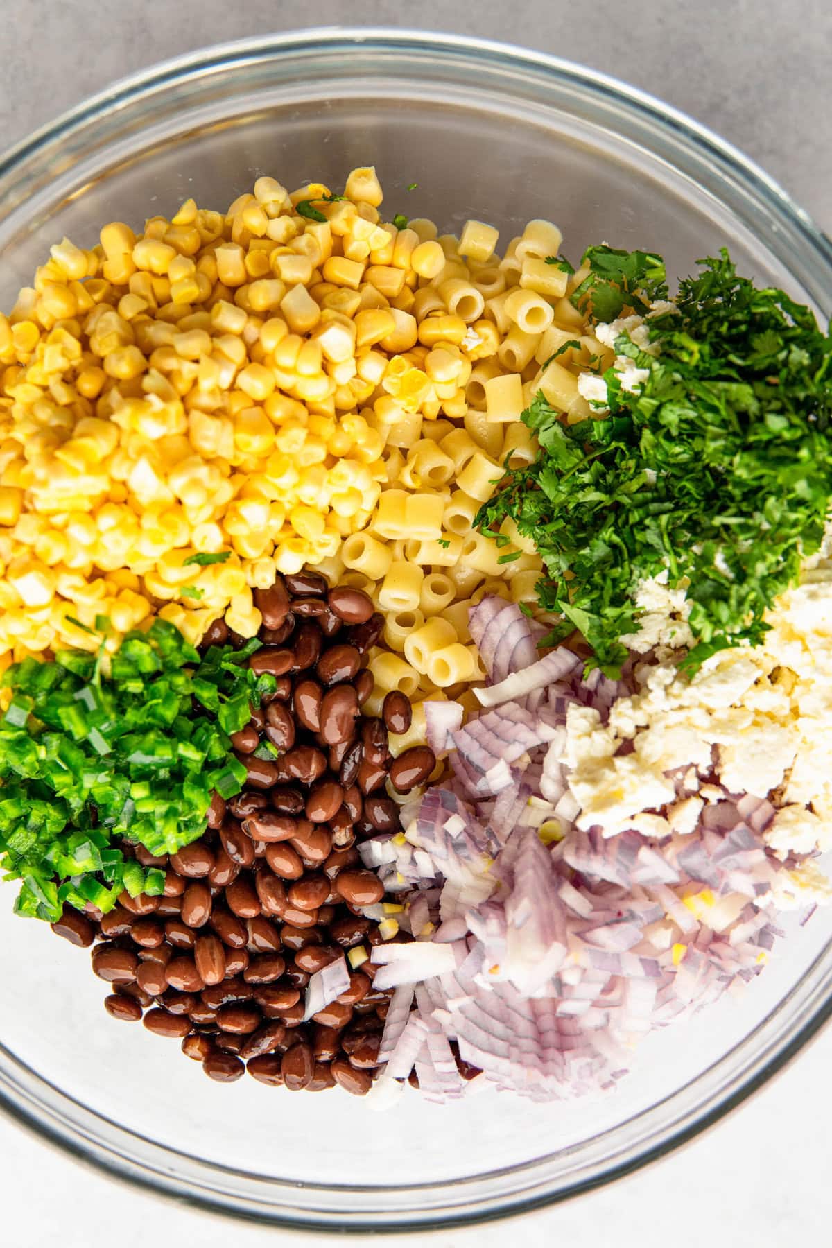 A clear glass bowl containing chopped parsley, diced red onion, black beans, corn kernels, and small pasta pieces.