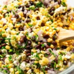 A bowl of corn pasta salad mixed with black beans, red onions, cilantro, and feta cheese. A wooden spoon is used for stirring.