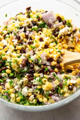 A bowl of corn pasta salad mixed with black beans, red onions, cilantro, and feta cheese. A wooden spoon is used for stirring.