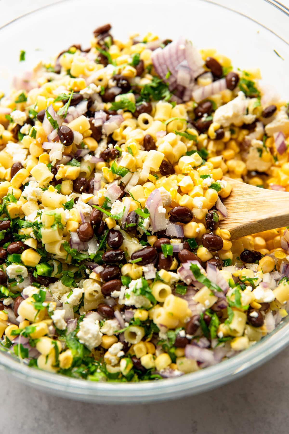 A bowl of corn pasta salad mixed with black beans, red onions, cilantro, and feta cheese. A wooden spoon is used for stirring.