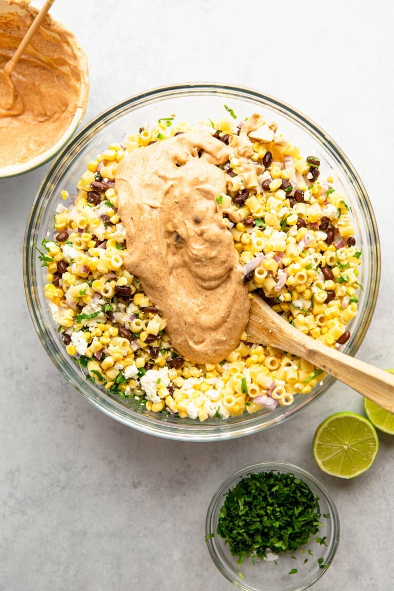 A glass bowl filled with a corn, black bean, pasta, and feta salad being mixed with a creamy dressing. There is a wooden spoon in the bowl, with a nearby bowl of dressing, lime halves, and chopped herbs.