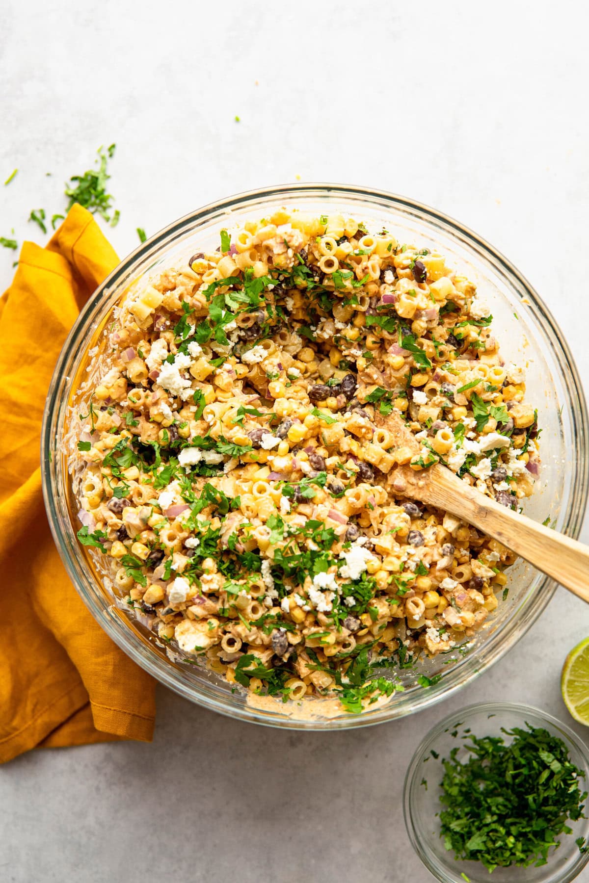 A glass bowl filled with pasta salad containing corn, black beans, red onions, and fresh herbs. A wooden spoon is in the bowl. An orange napkin is placed beside the bowl. A small bowl of herbs is nearby.