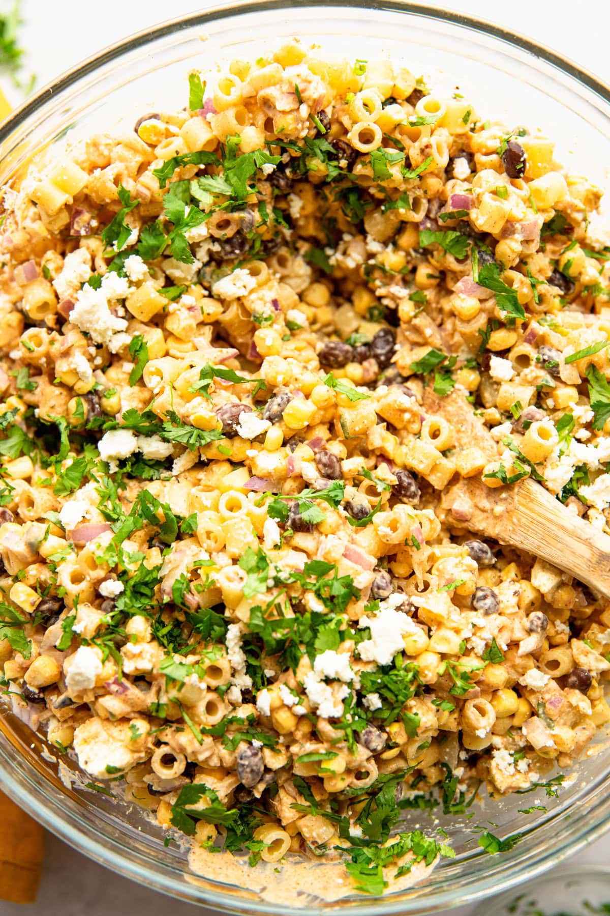 A glass bowl filled with a mixed pasta salad that includes elements like corn, black beans, chopped vegetables, crumbled cheese, and garnished with fresh herbs. A wooden spoon is partially visible on the right.