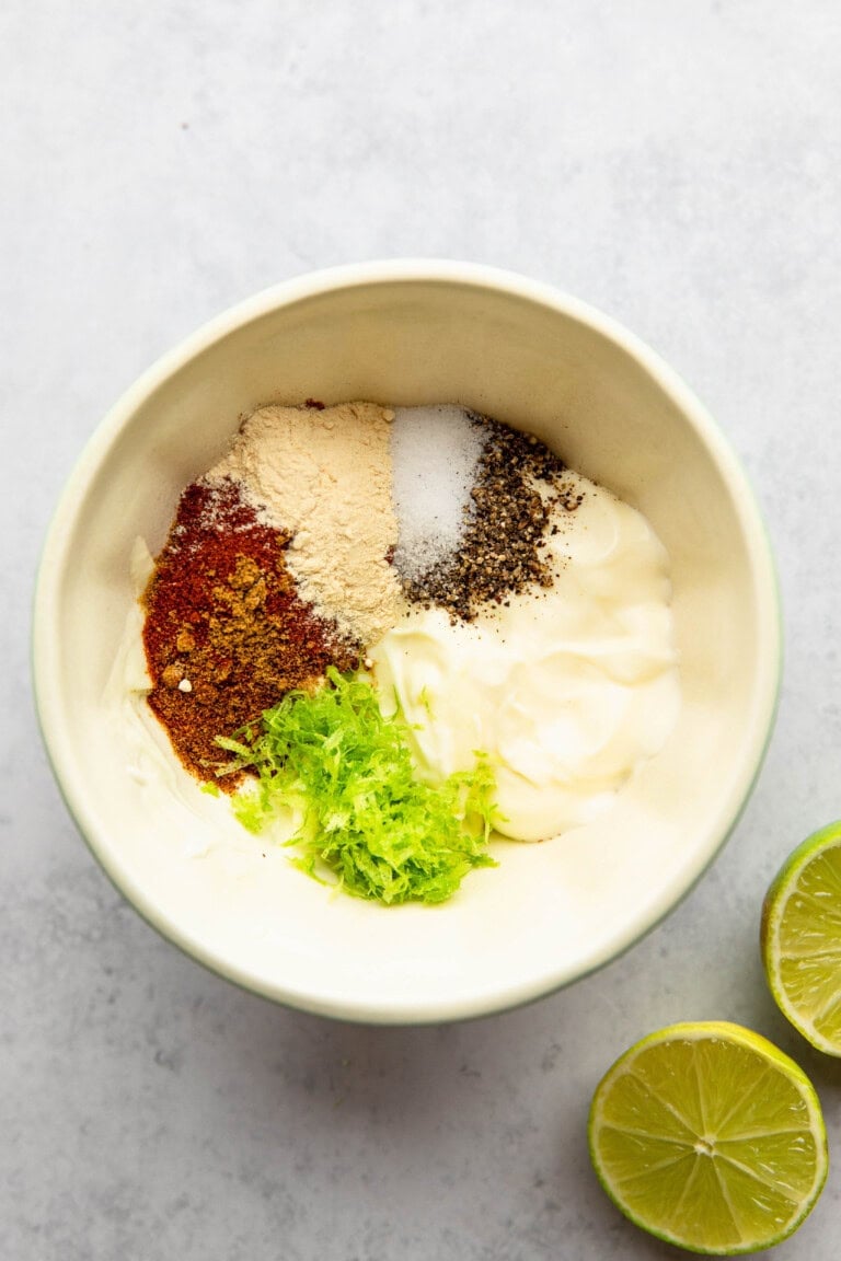 A bowl containing mayonnaise, ginger powder, salt, pepper, paprika, dried herbs, and lime zest, with half-cut limes on the side.