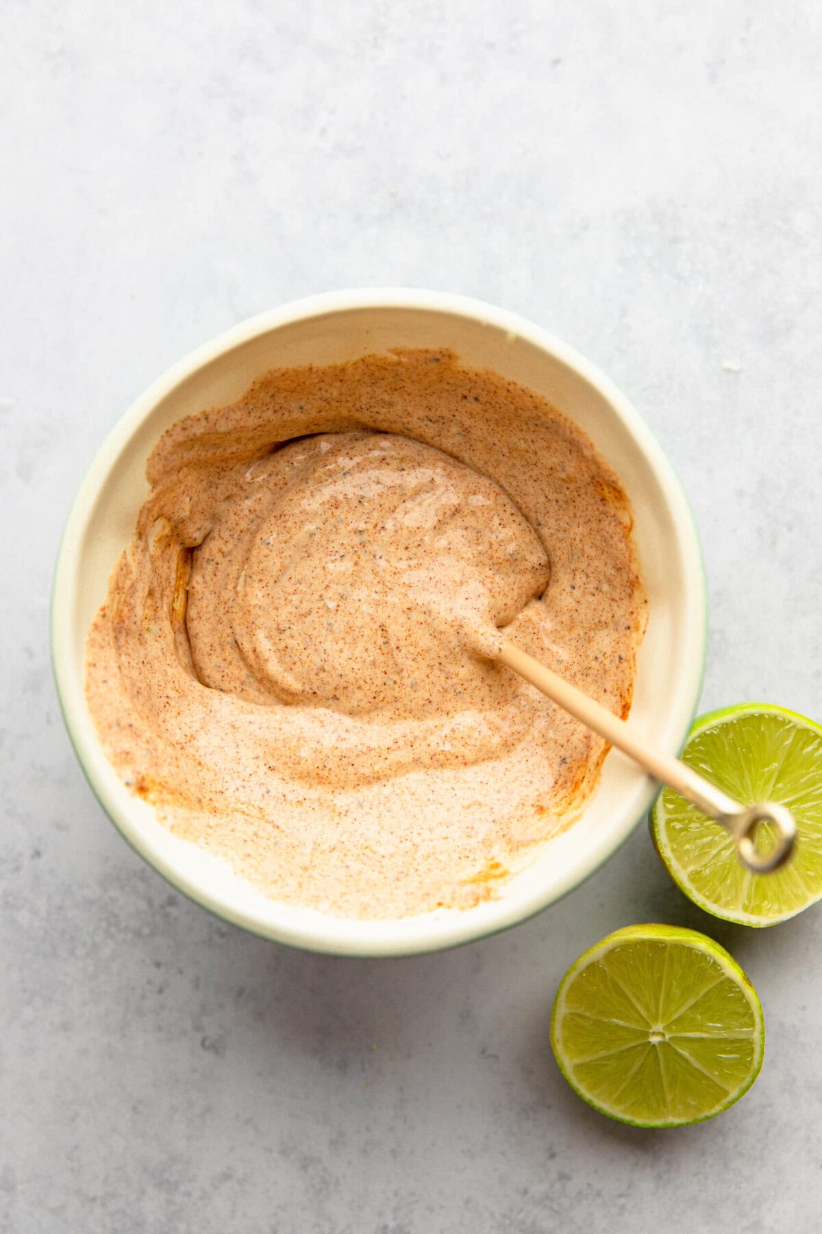 A bowl filled with a creamy, beige dipping sauce with visible spices, accompanied by a small stir stick. Two lime halves are placed next to the bowl on a light gray surface.