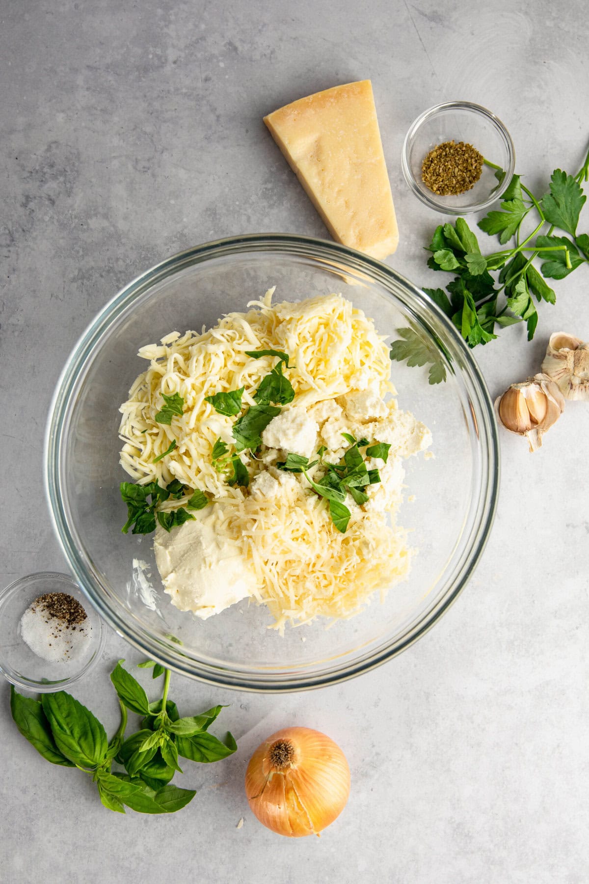 A glass bowl containing grated cheese, ricotta, and parsley. Surrounding the bowl are herbs, spices, a garlic bulb, a basil leaf, an onion, and a block of cheese on a grey countertop.