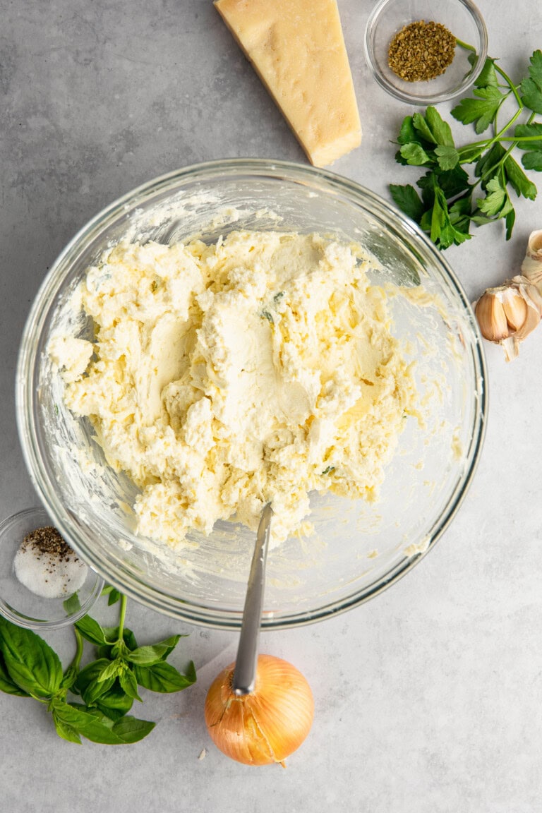 A mixing bowl filled with a thick, creamy mixture is surrounded by a block of cheese, fresh parsley, garlic bulbs, an onion, fresh basil leaves, salt, and dried herbs on a gray countertop.