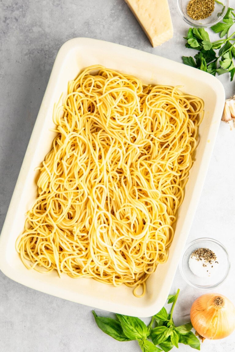 A tray of cooked spaghetti is surrounded by ingredients including parmesan cheese, basil, garlic, pepper, and an onion on a marble surface.