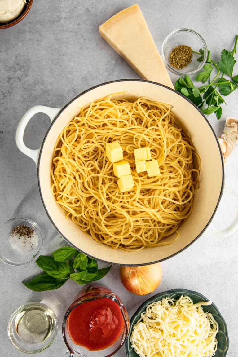 A pot of cooked spaghetti with butter cubes on top, surrounded by ingredients including a wedge of cheese, herbs, garlic, an onion, a bowl of shredded cheese, a bowl of sauce, and small dishes of spices.