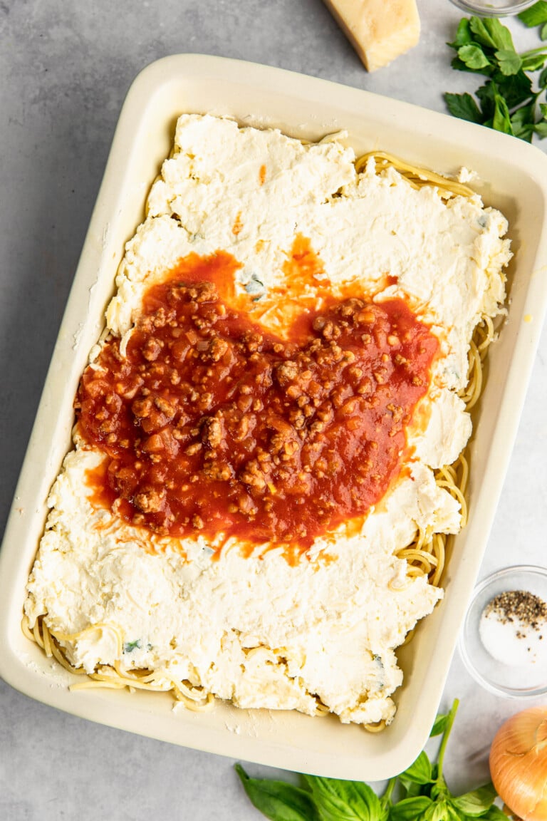 A baking dish filled with uncooked lasagna layers, ricotta cheese, and a portion of meat sauce. Ingredients like basil, Parmesan cheese, onion, and spices are nearby.