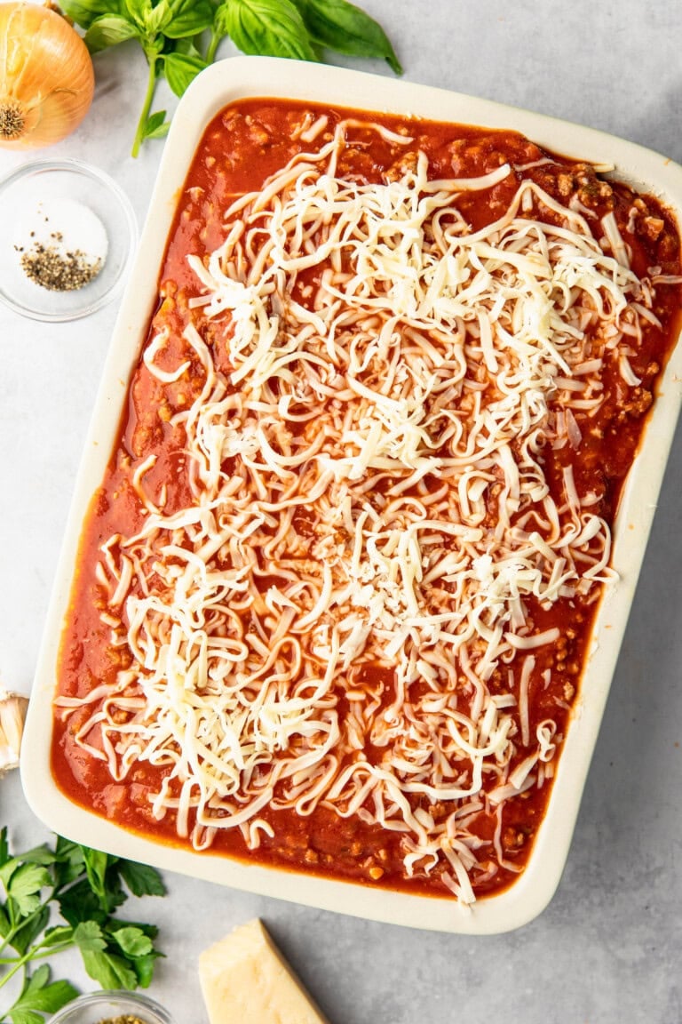 A rectangular baking dish filled with lasagna, topped with tomato sauce and shredded cheese, sits on a countertop. Fresh basil, onion, a dish of pepper, and a wedge of cheese are nearby.