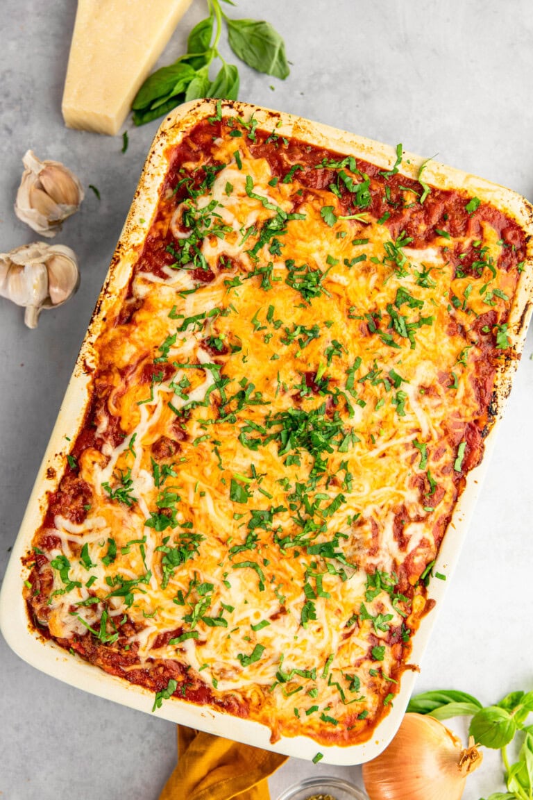 A baked dish of vegetable lasagna topped with melted cheese and fresh herbs, placed on a light grey surface with garlic cloves, a block of cheese, and fresh basil on the side.