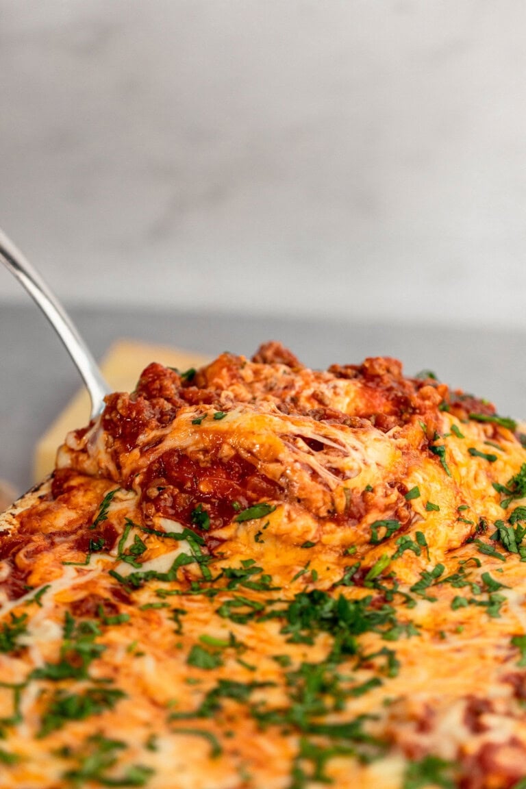 A close-up of a freshly baked lasagna slice being lifted from a dish, showcasing layers of melted cheese, rich tomato sauce, ground meat, and herbs.