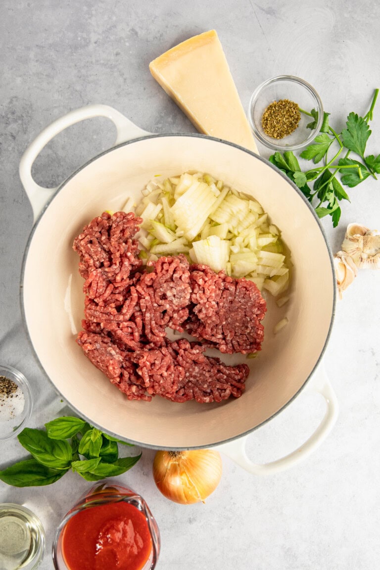 White pot with raw ground meat and chopped onions. Surrounding items include cheese wedge, seasoning in a bowl, parsley, garlic, fresh basil, jar of tomato sauce, whole onion, and glass of oil.