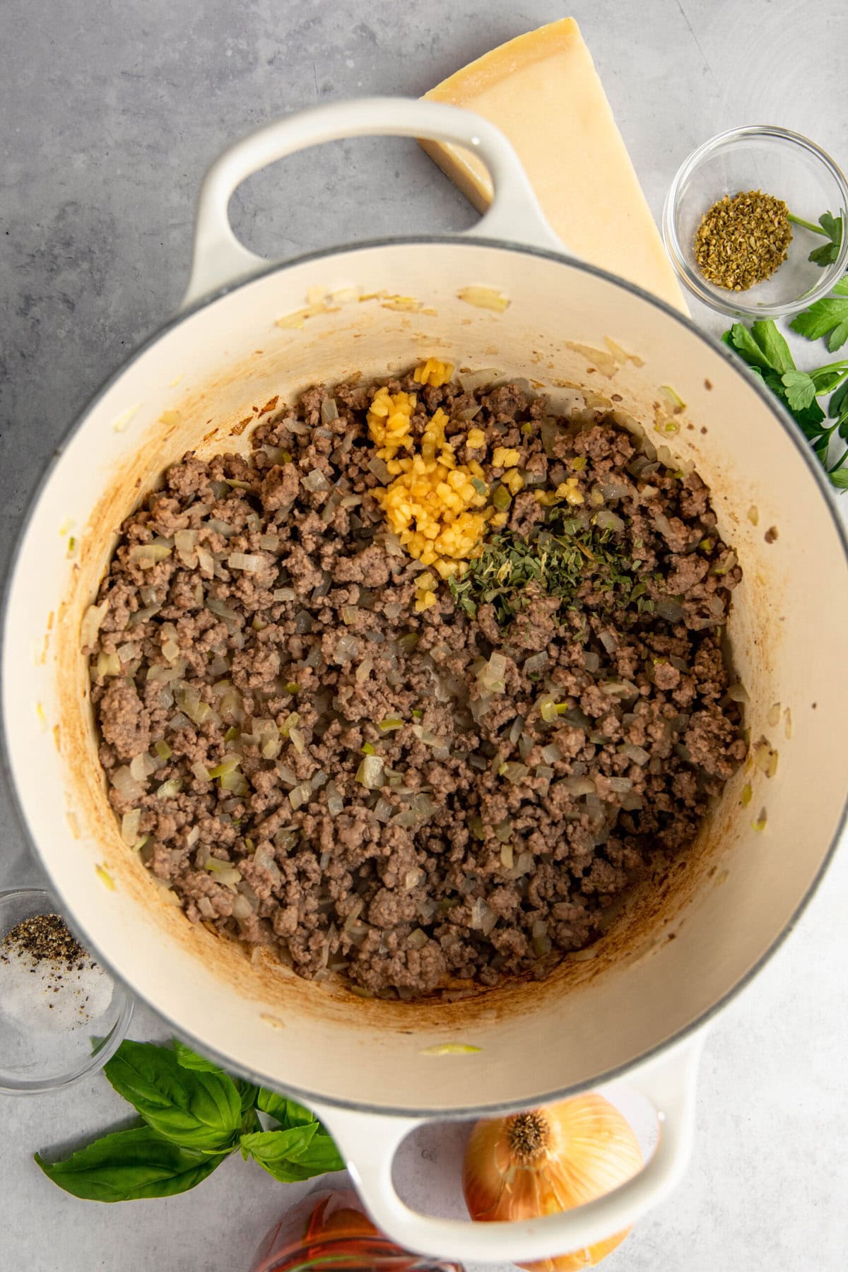 A pot containing cooked minced meat mixed with diced onions and garlic, placed on a gray surface with additional herbs and ingredients surrounding it.