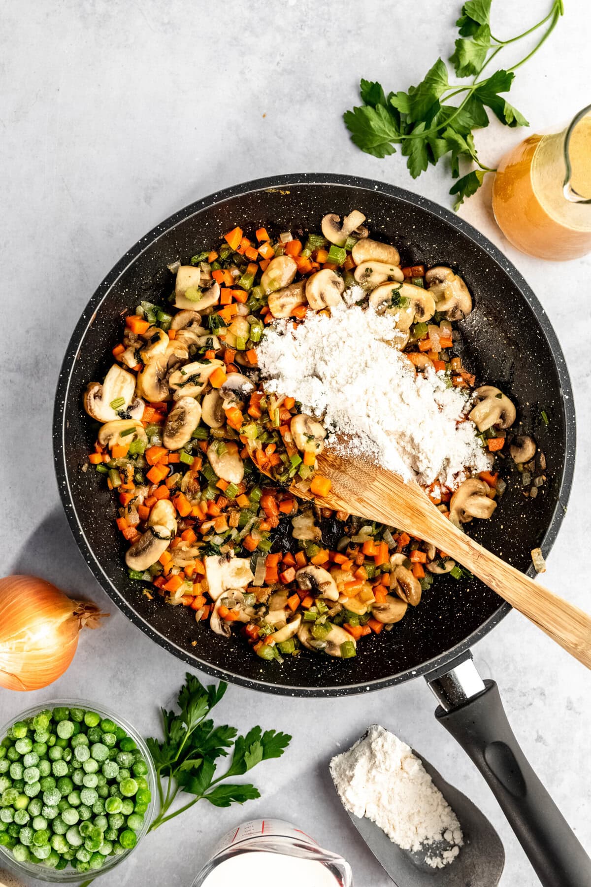 A frying pan on a grey surface holds a mixture of chopped vegetables and mushrooms with a pile of flour. Surrounding the pan are fresh parsley, an onion, frozen peas, flour, and a glass container of liquid.