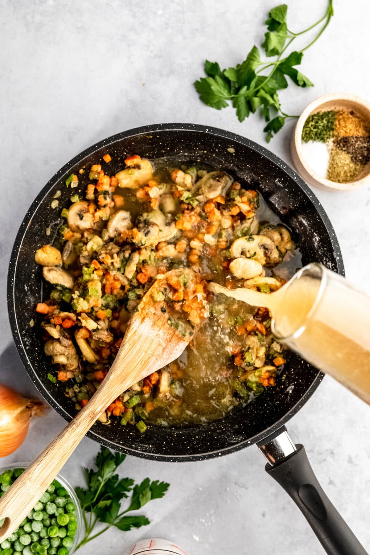 A wooden spoon stirs a mixture of vegetables in a black pan while broth is being poured in. A bowl of spices, chopped parsley, and an onion are nearby on a light gray surface.