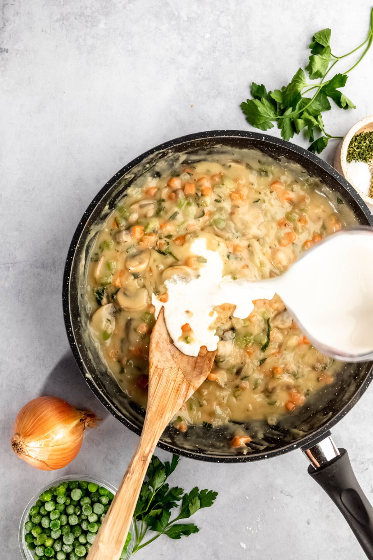 A wooden spoon stirs a creamy vegetable mixture in a pan as milk is being poured in, with an onion, peas, parsley, and dried herbs placed around the pan.