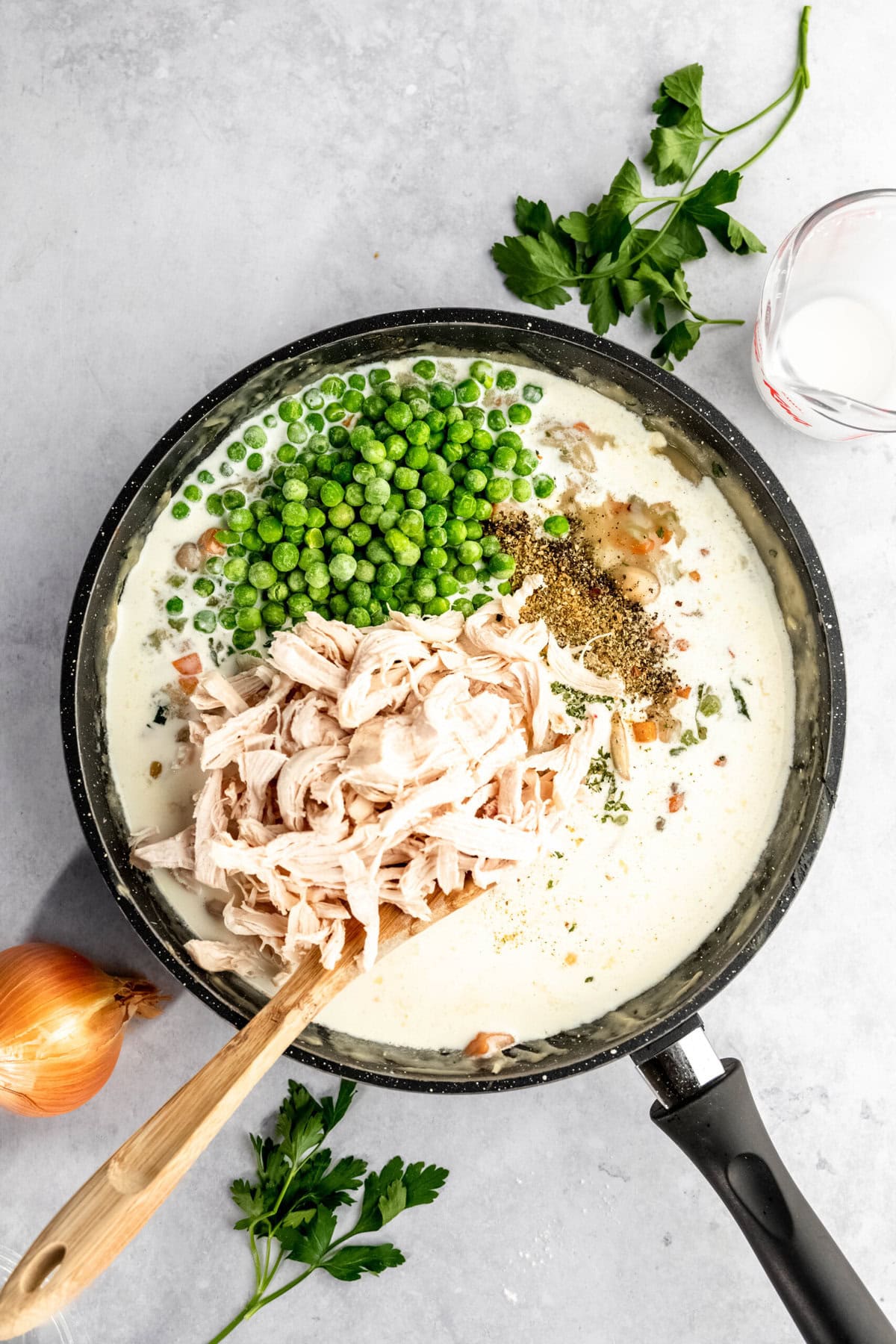 A skillet filled with a creamy sauce, shredded chicken, peas, and seasonings is being stirred with a wooden spoon. Fresh parsley, an onion, and a glass of milk are on the countertop beside it.