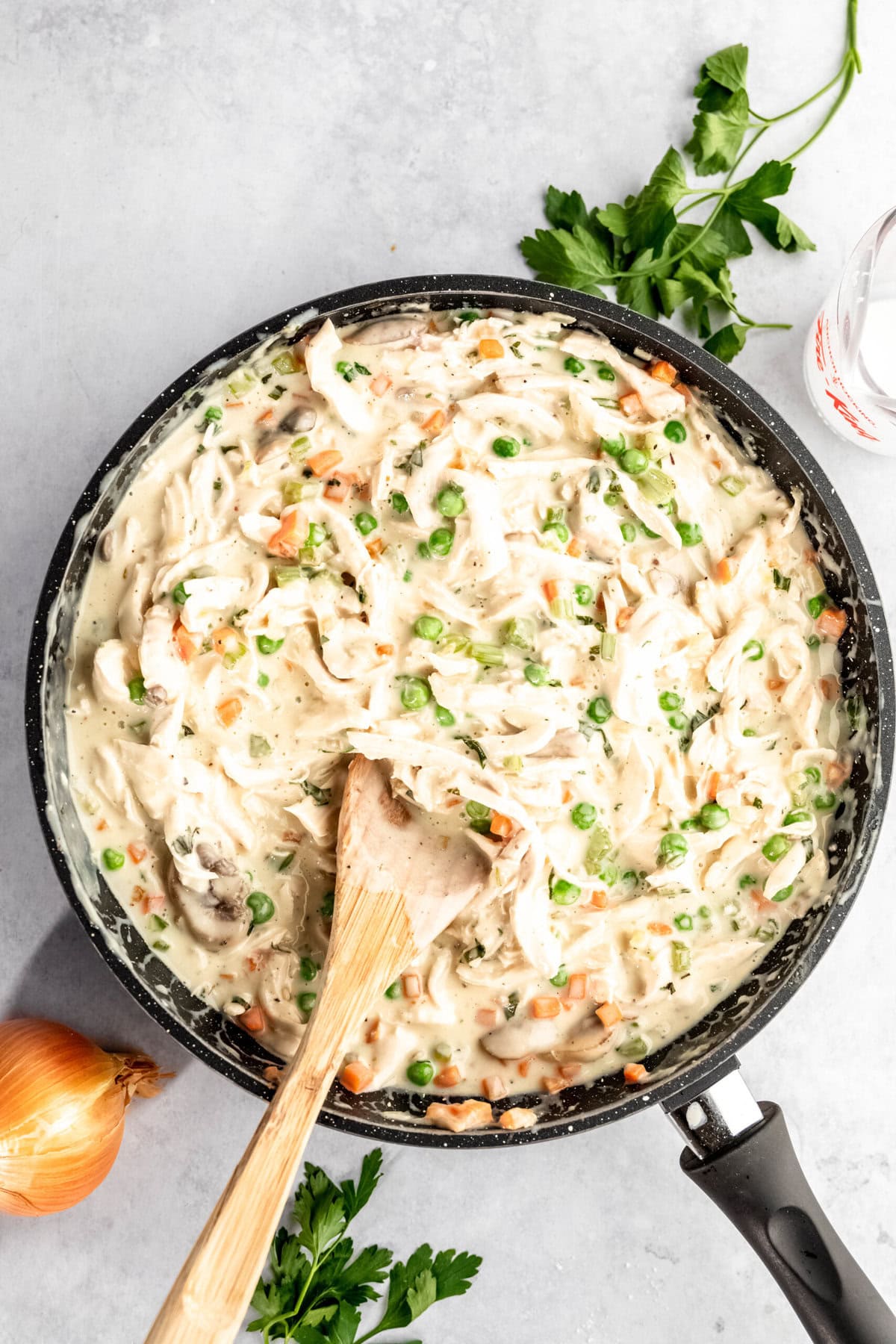 A skillet containing creamy chicken pot pie filling with shredded chicken, peas, carrots, and a wooden spoon. Fresh parsley and an onion are placed next to the skillet.