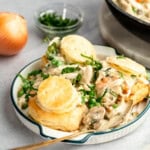 A plate of creamy chicken and vegetables topped with biscuits, with a wooden spoon, an onion, a bowl of chopped herbs, and a skillet in the background.