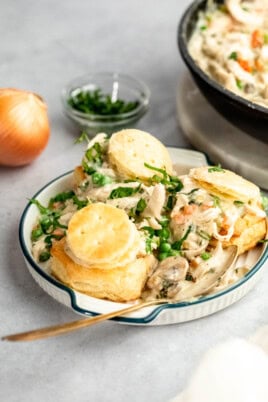 A plate of creamy chicken and vegetables topped with biscuits, with a wooden spoon, an onion, a bowl of chopped herbs, and a skillet in the background.