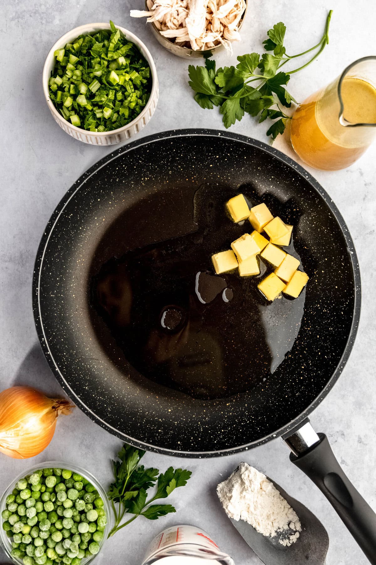 A frying pan with butter and oil is placed on a countertop surrounded by bowls of chopped greens, peas, parsley, a whole onion, shredded chicken, broth, and flour.