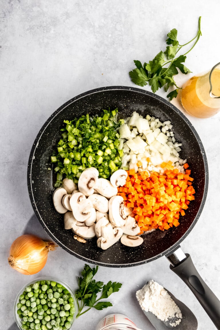 A skillet contains chopped green bell peppers, onions, carrots, and sliced mushrooms. Surrounding items include peas, an onion, fresh parsley, a small dish of flour, and a jug of liquid.