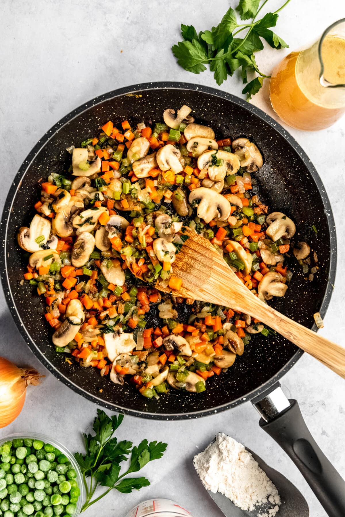 A skillet filled with diced vegetables including mushrooms, carrots, and peas being sautéed with a wooden spatula. Surrounding the skillet are ingredients such as parsley, flour, an onion, peas, and broth.