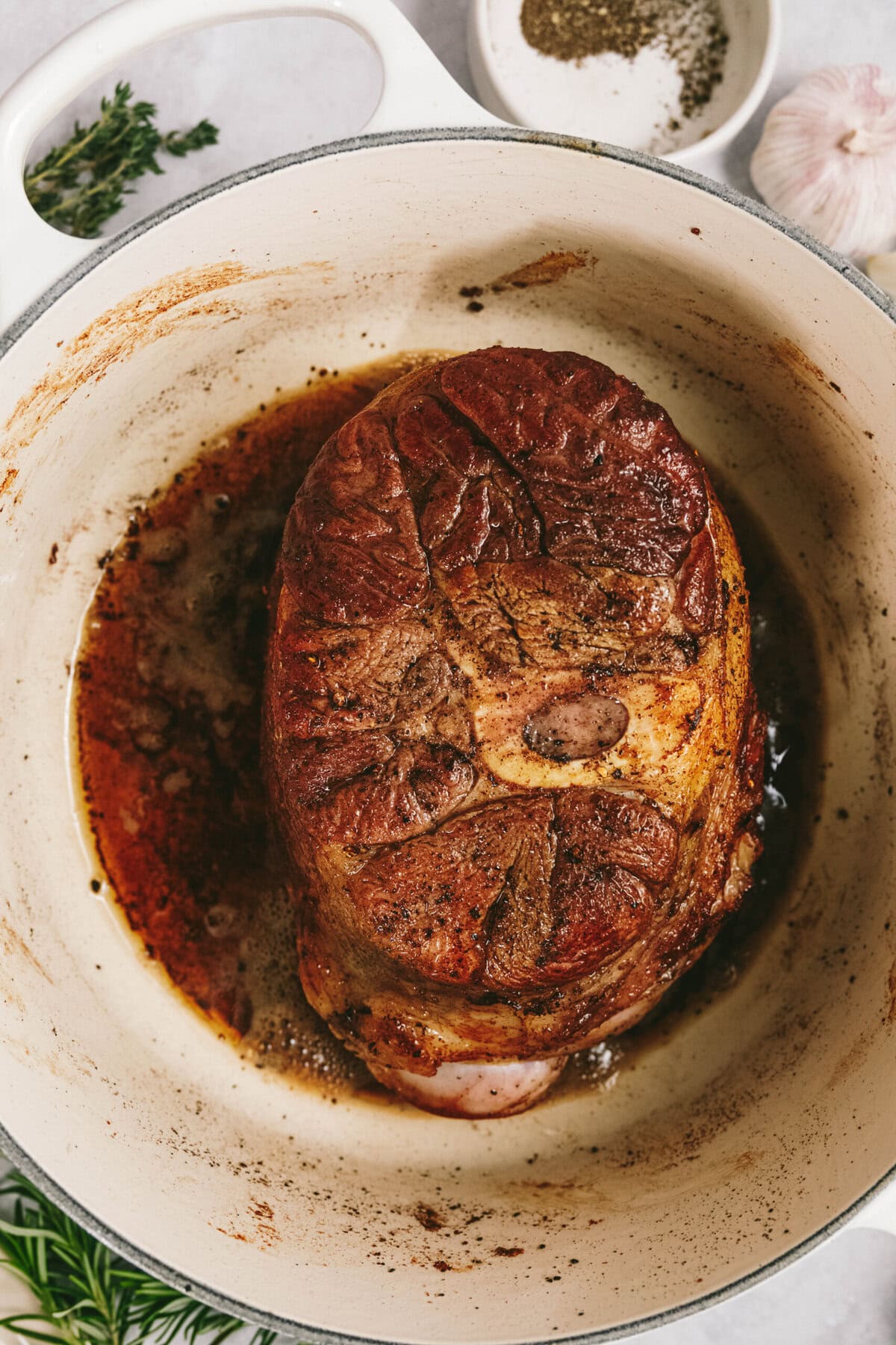 An arm roast is browning beautifully in a white cast iron pot. Fresh herbs, a whole garlic bulb, and a bowl of pepper are visible in the background.