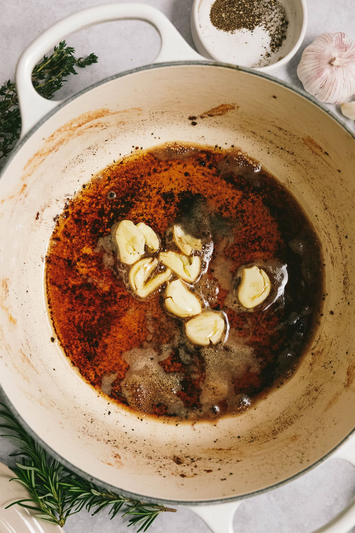 A white pot contains melted butter and browned bits with garlic cloves heating in it, perfect for an arm roast. Herbs, garlic, and pepper are visible around the pot.