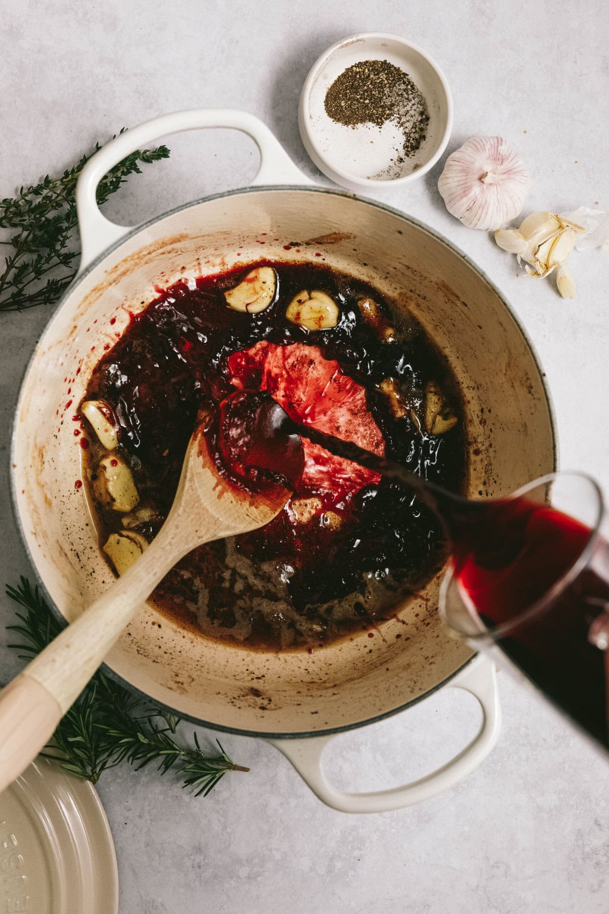 A white pot filled with simmering dark liquid, pieces of garlic, and a succulent arm roast in the center. A wooden spoon is stirring the contents while red wine is being poured in from a measuring cup.