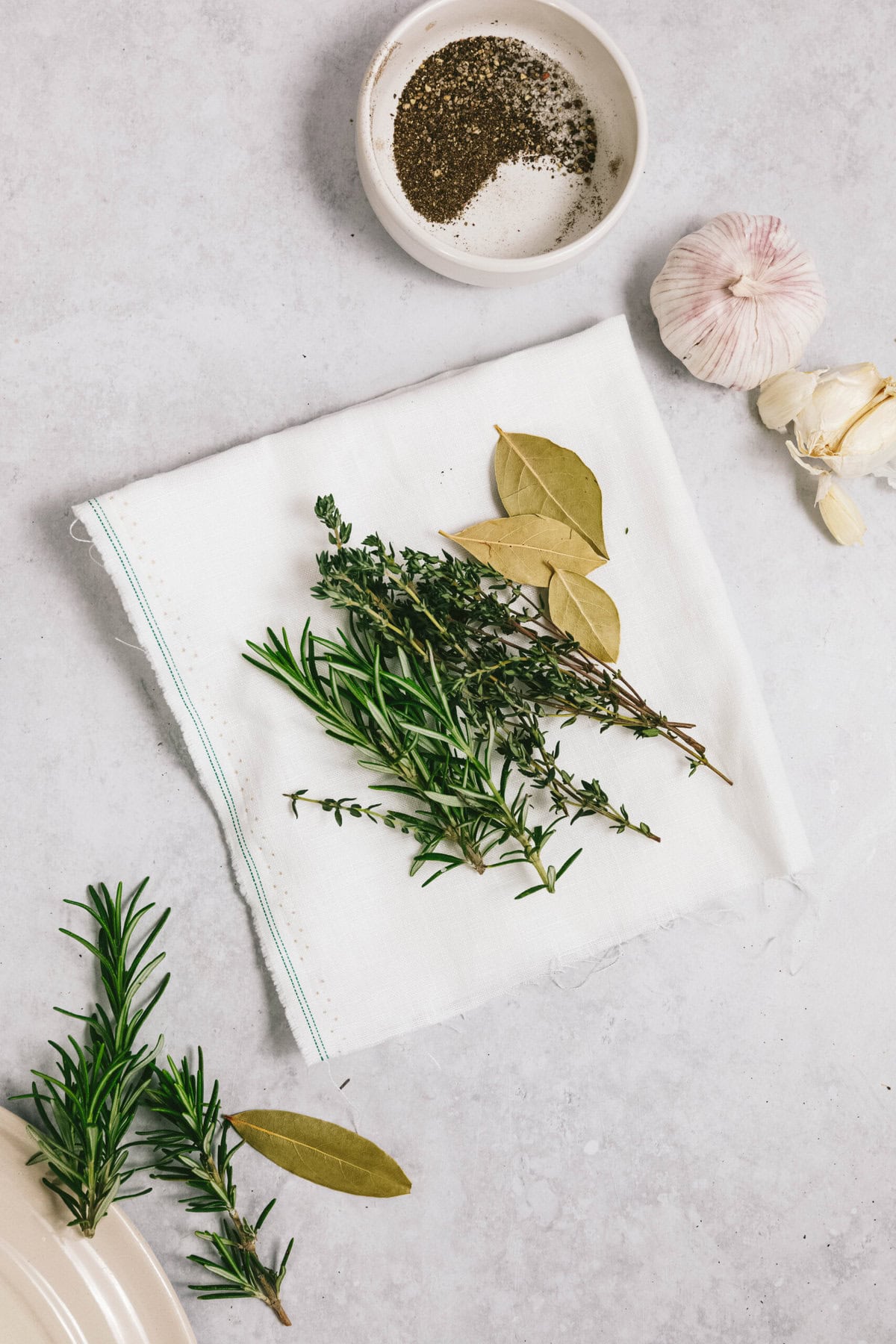 A white cloth with herbs including rosemary, thyme, and bay leaves rests beside an arm roast. To the side are additional rosemary sprigs, a small bowl of ground pepper, and garlic cloves on a light-colored surface.