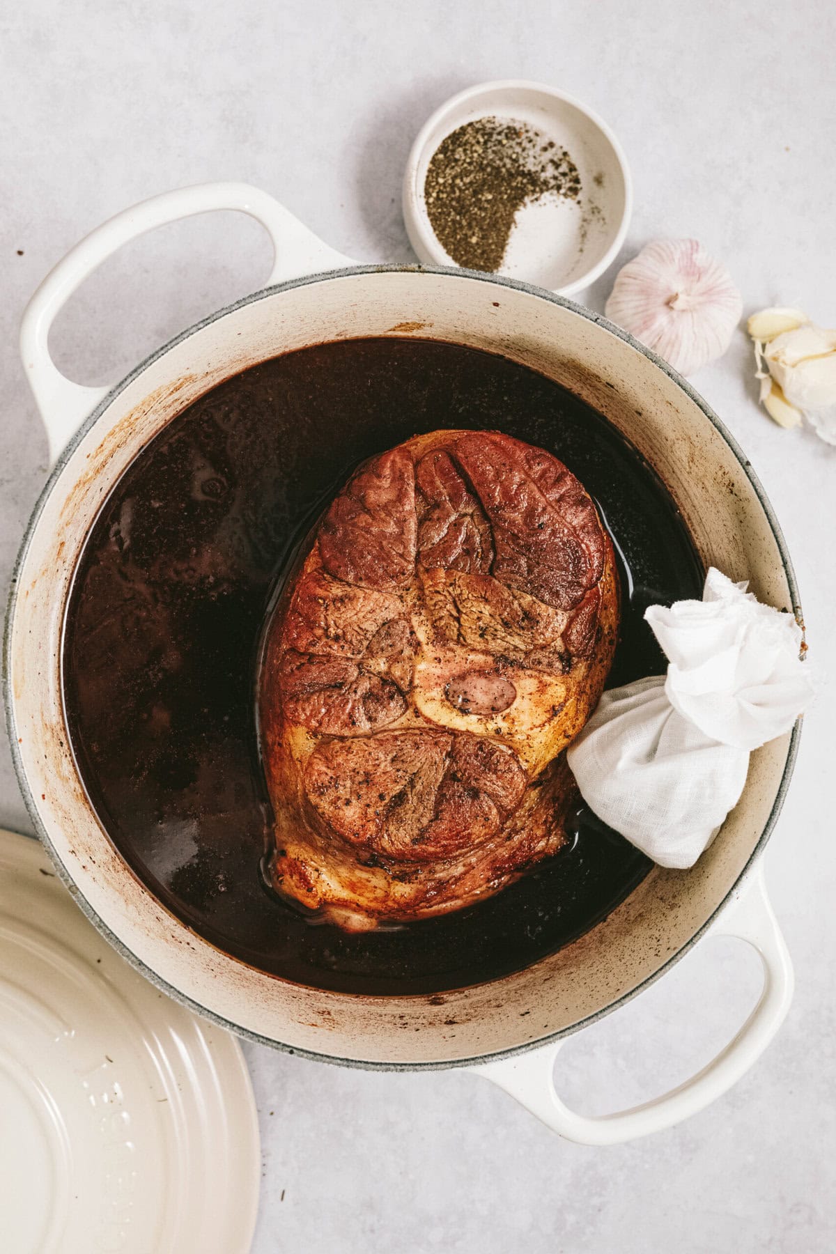 A cooked arm roast in a white Dutch oven filled with dark broth, with a seasoning bowl, garlic cloves, and a lid nearby.