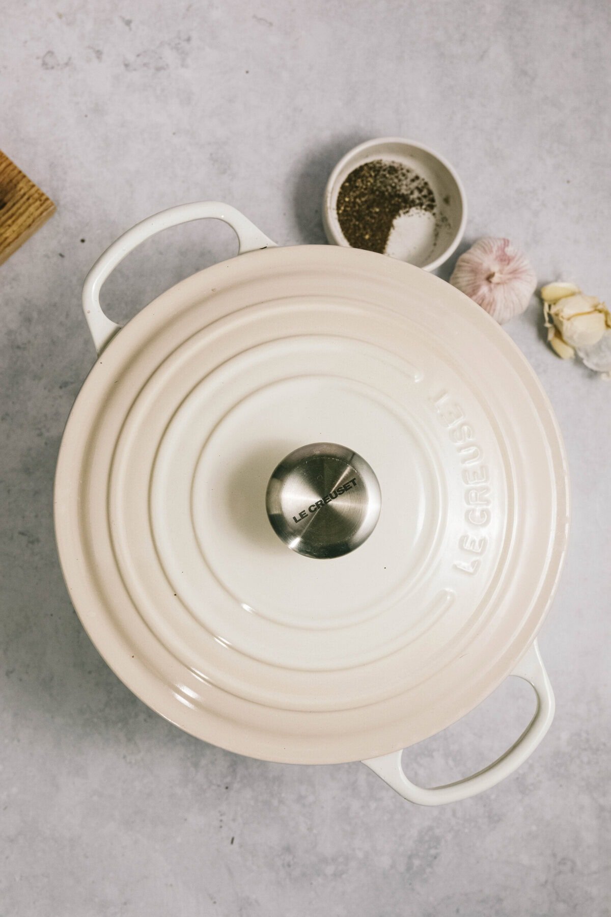 A white Le Creuset dutch oven with a closed lid sits on a countertop, next to a small bowl of salt and pepper, a garlic bulb, and a few garlic cloves—perfect for preparing that tender arm roast.