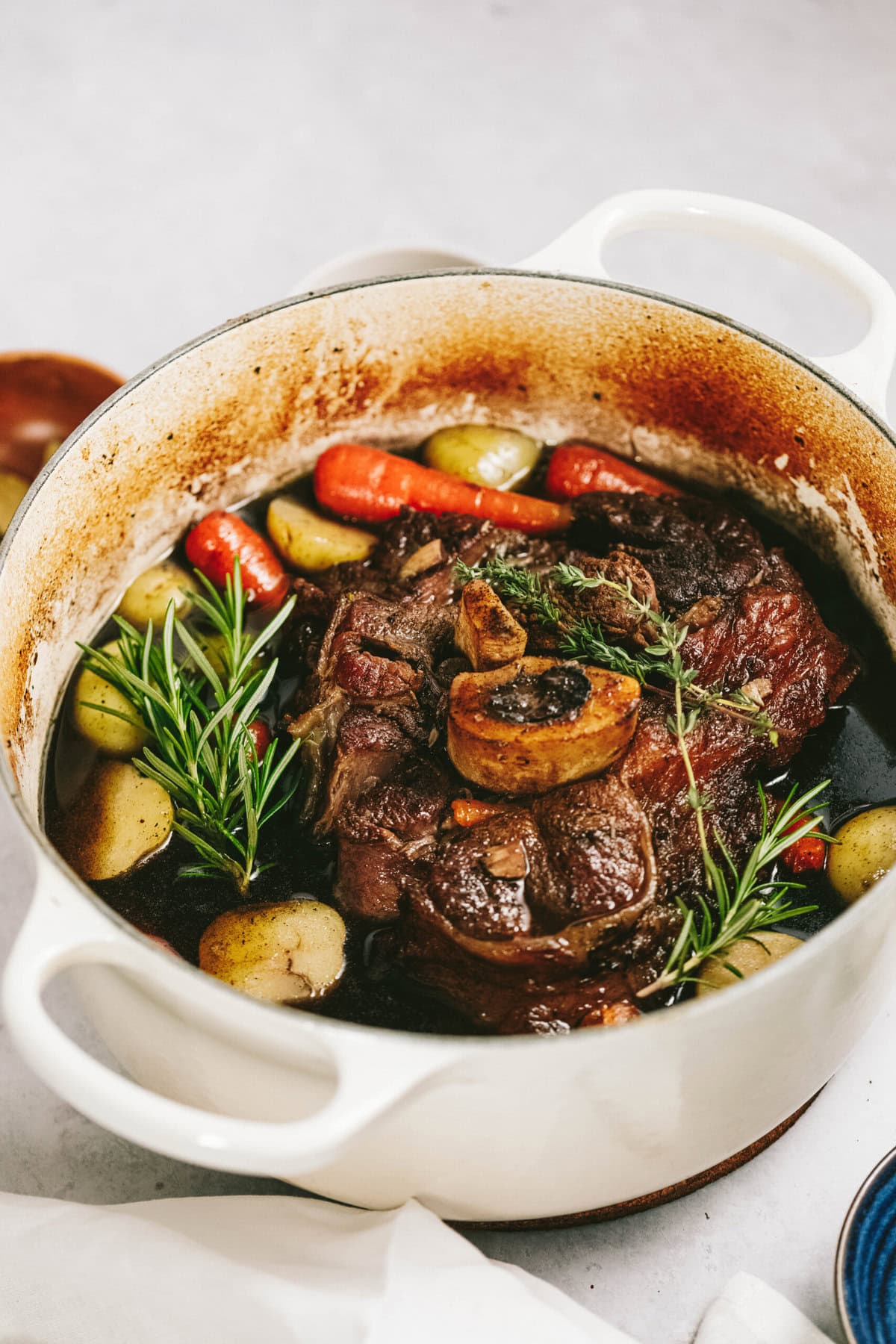 A white pot filled with cooked osso buco, featuring braised arm roast, bone marrow, potatoes, carrots, and garnished with fresh rosemary.