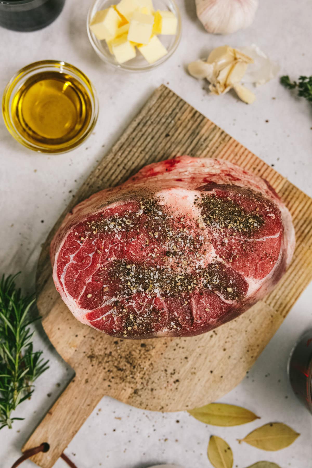 Raw seasoned arm roast on a wooden cutting board surrounded by ingredients including olive oil, butter, garlic, herbs, and bay leaves.