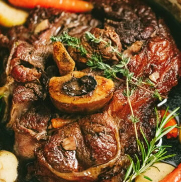 A close-up of an arm roast with bone-in beef surrounded by cooked carrots, potatoes, and herbs in a pot.