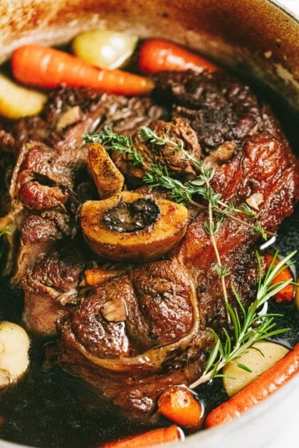 A close-up of an arm roast with bone-in beef surrounded by cooked carrots, potatoes, and herbs in a pot.