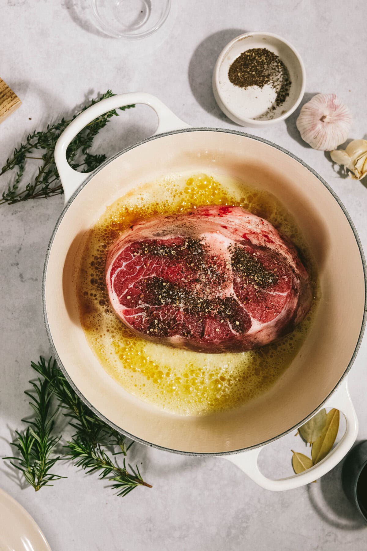 A large arm roast is being seared in a pot with butter, surrounded by various herbs and spices.