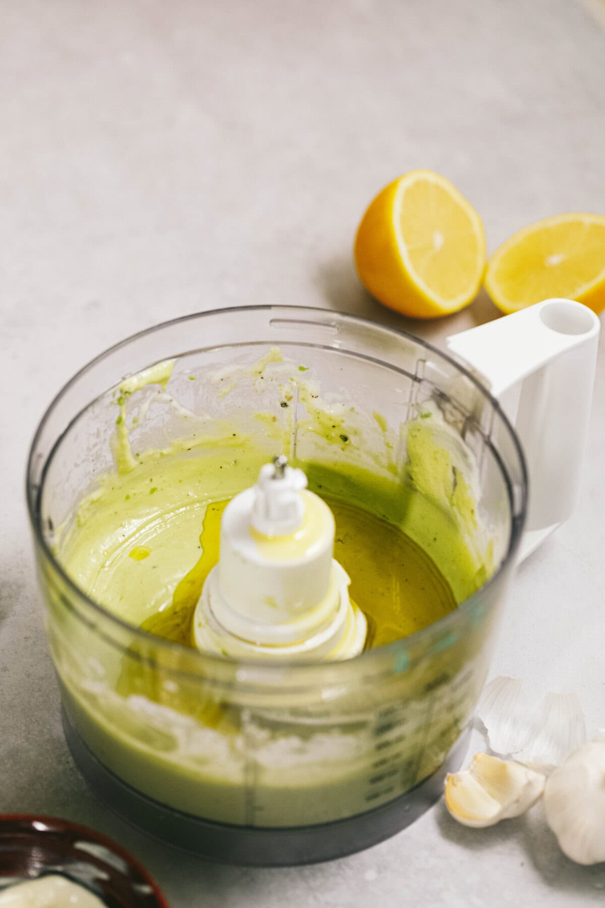 A food processor with green puree sits on a countertop. A halved lemon and garlic cloves are nearby.