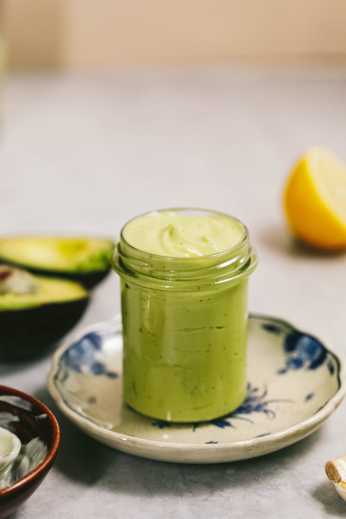 A jar of creamy green sauce sits on a small plate. Halb of an avocado and a lemon wedge can be seen in the background.