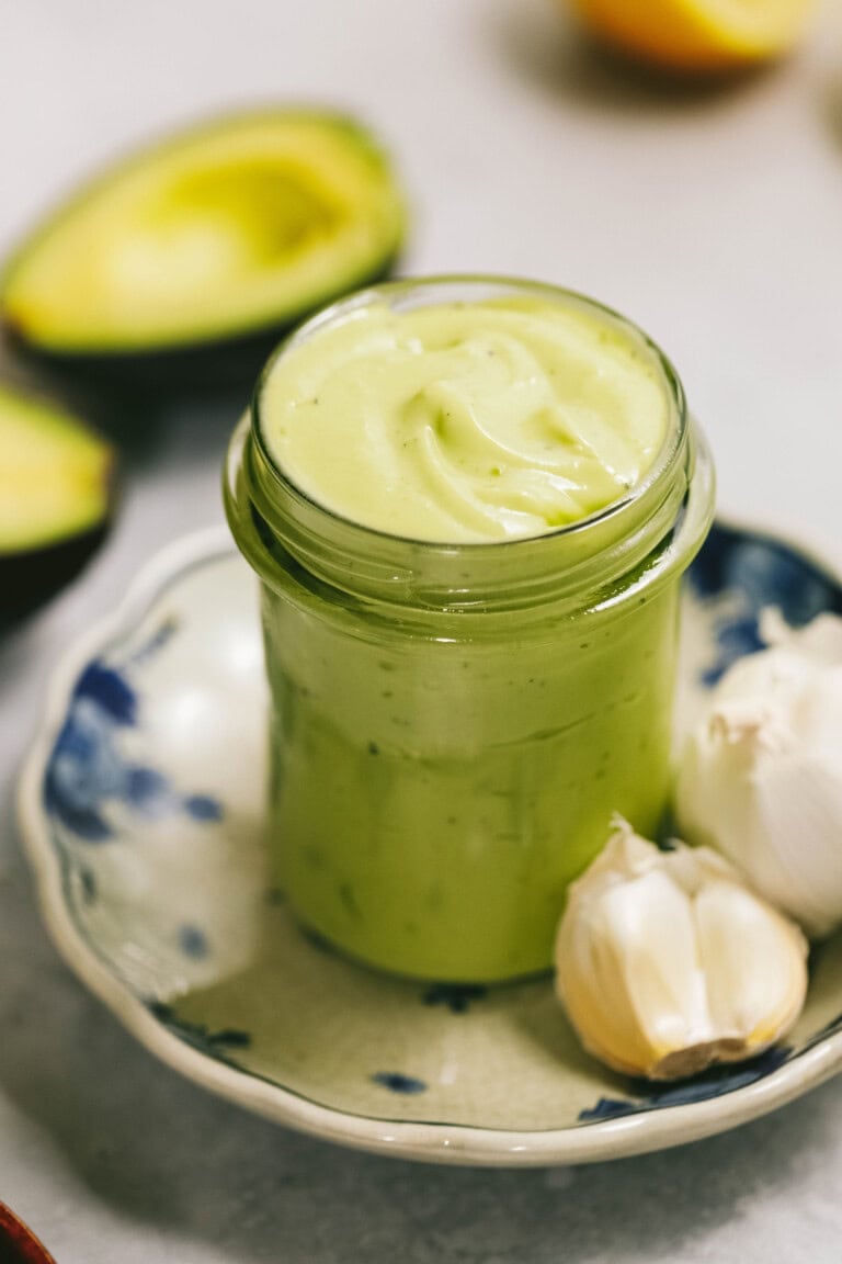 A glass jar filled with creamy green avocado sauce is placed on a ceramic dish beside two garlic cloves and a halved avocado.